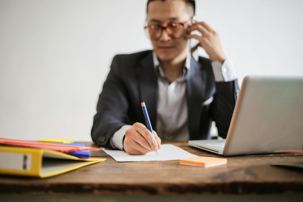 pexels-photo-3760612-3760612 Businessman talking on phone while writing notes and using a laptop at desk.